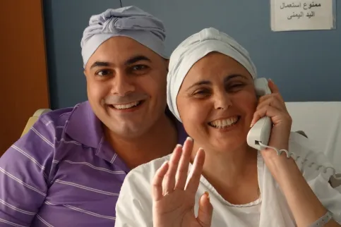 Smiling man wearing a purple t-shirt and headscarf with smiling woman with cancer on the phone, a hand raised, also with a headscarf