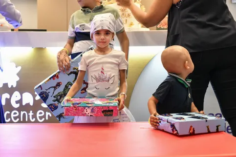 A child wearing a headscarf facing the camera is opening a present, while another present on the table is in front of a younger child with bald head, face turned away.