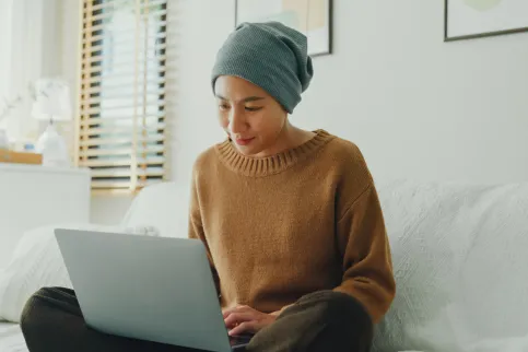 Young Asian woman wearing a headscarf comfortably working on her laptop, seated on a sofa in a well-lit, plant-decorated room.