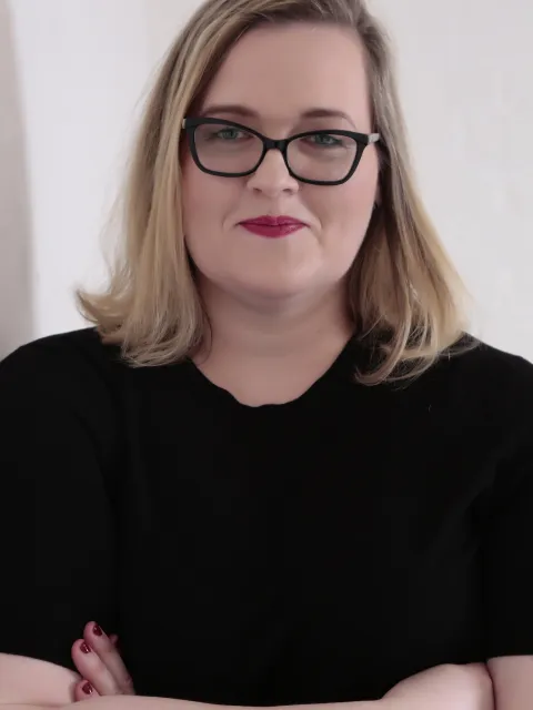 Headshot of blond Caucasian woman wearing black glasses and black shirt with arms crossed